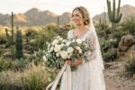 A bride holding a lush cascade bouquet of white roses and desert greenery.